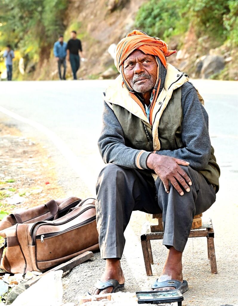 "An elderly man wearing an orange turban and a gray jacket, sitting by the roadside with his belongings in a village near the Tashita Institute in Dharamshala."